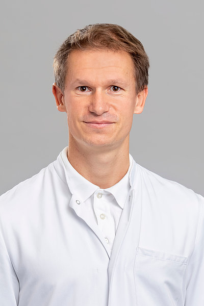 Mann mit kurzem Haar trägt weißen Kittel und weißes Hemd vor grauem Hintergrund